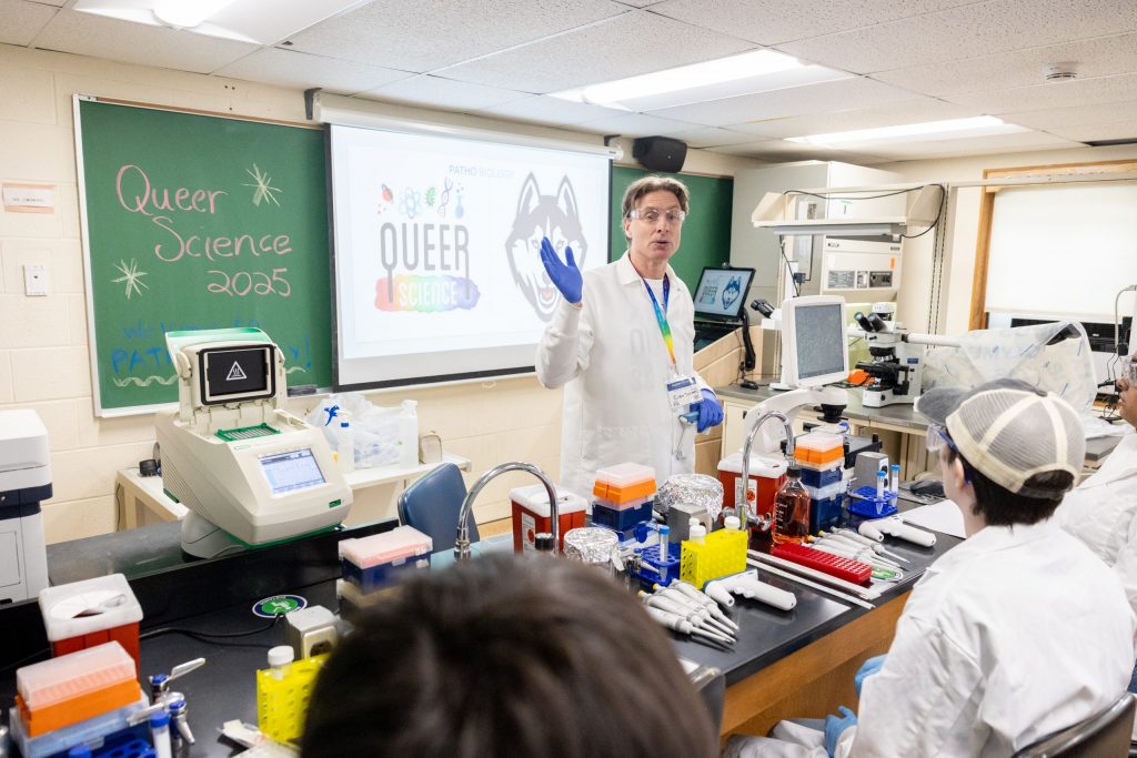 Edan Tulman, instructor in residence of pathobiology at UConn, stands in front of a green chalkboard with a projected presentation to lead a pipette tutorial in Atwater Building during the Queer Science Conference on Sunday, June 8, 2025. (Sydney Herdle/UConn Photo)