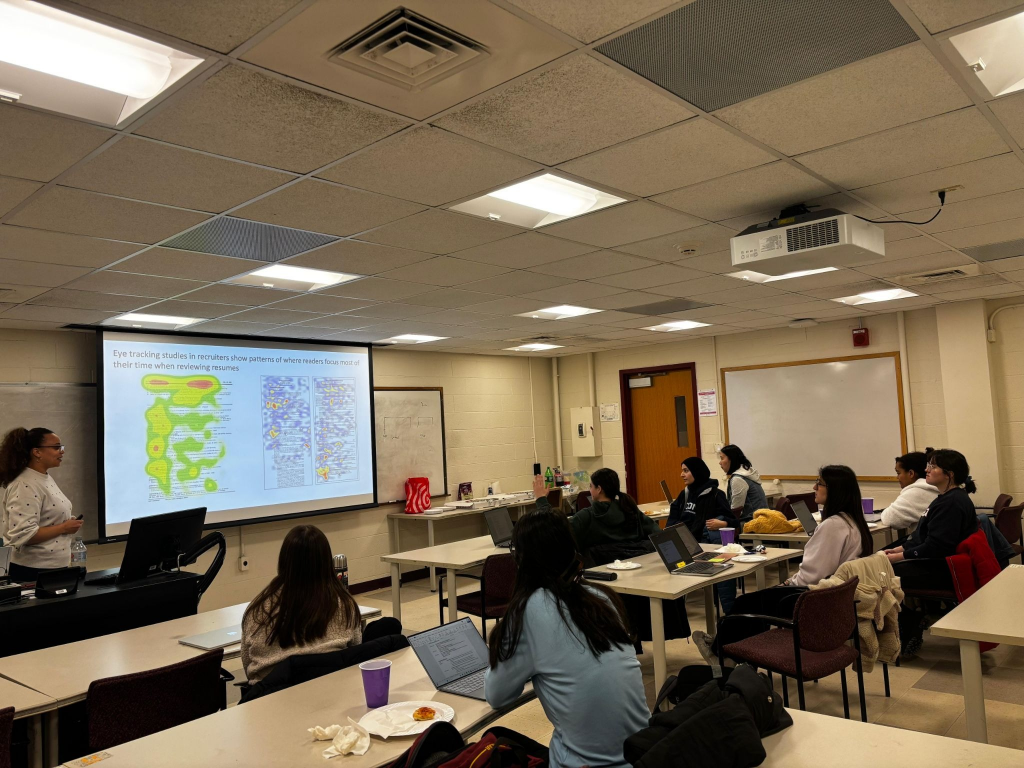 Grad SWE members sitting in a classroom watching a projected presentation about resumes, with an example resume with a heatmap of most veiwed areas on the board.