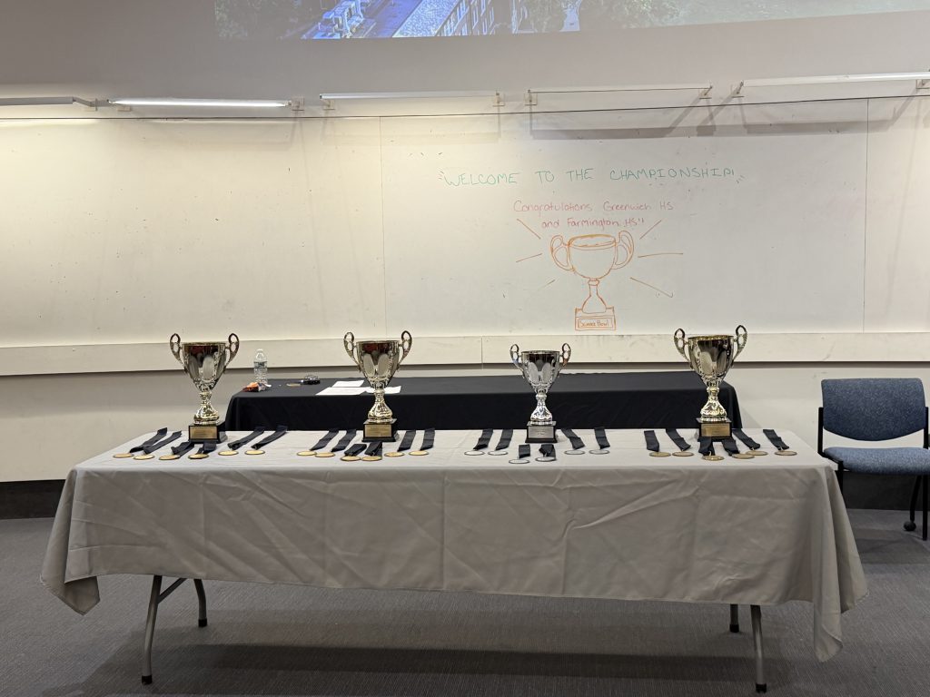 The trophies and medals for the winners and runner-ups of the Connecticut and UConn House science bowl tournaments, with a drawing on the whiteboard in the background of a trophy.