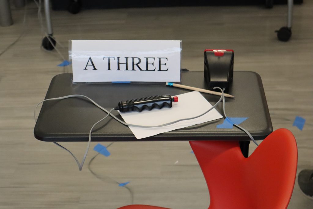 A close-up image of the desk setup used by students at the Connecticut Regional Science Bowl, featuring the "A THREE" player tag, handheld buzzer and light, a sheet of paper, and a pencil on a black desk with a red chair.