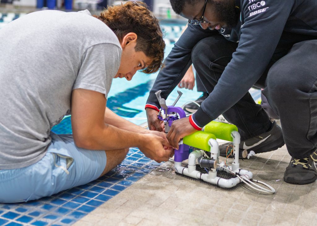 Two students sitting on the ground at the edge of an athletics swimming pool, making modifications to a small underwater robot.