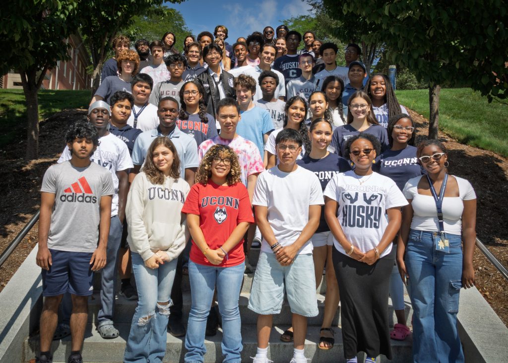 A group of diverse students wearing UConn apparel taking a photo on the Northwest Dorm stairs on a sunny day.