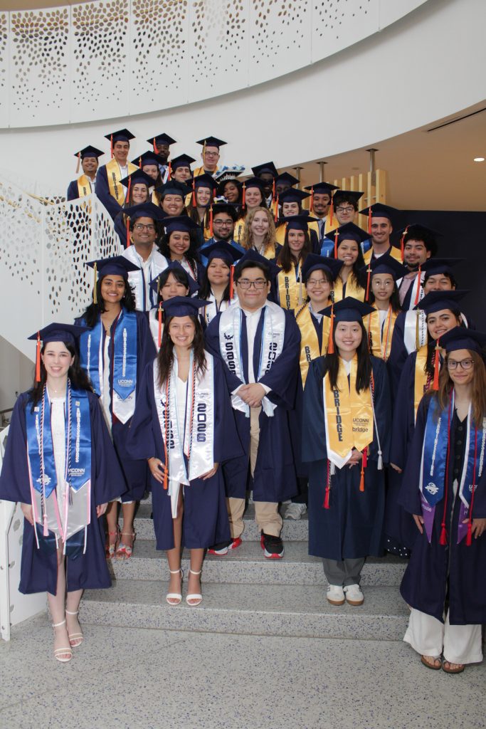 The VII 2026 graduate class gathered for a group photo on the staircase within the Gant Science Complex on the UConn Storrs campus. All of the students are wearing their caps and gowns, orange engineering tassles, and other graduation regalia.