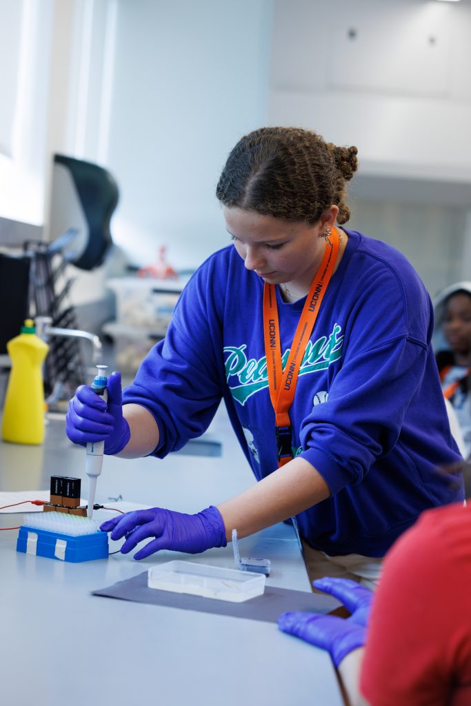 A female student wearing a blue long sleeve shirt and an orange College of Engineering layard uses scientific equipment to fill small vials.