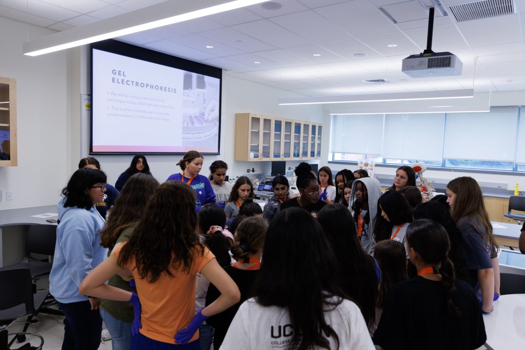 A group of students participating in the SPARK program gathered around an instructor in a classroom. The projector in the background introduces a lab technique "Gel Electrophoresis".