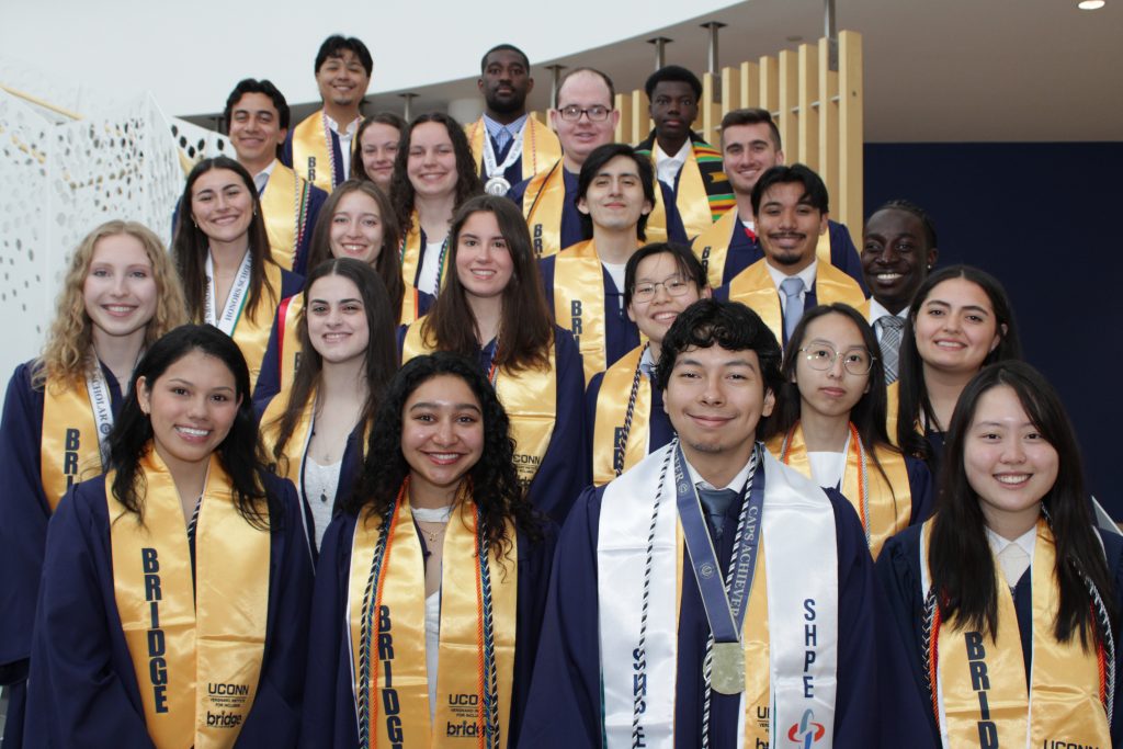 Members of the BRIDGE 2026 class taking a group photo wearing graduation regalia, including their matching yellow BRIDGE stoles.