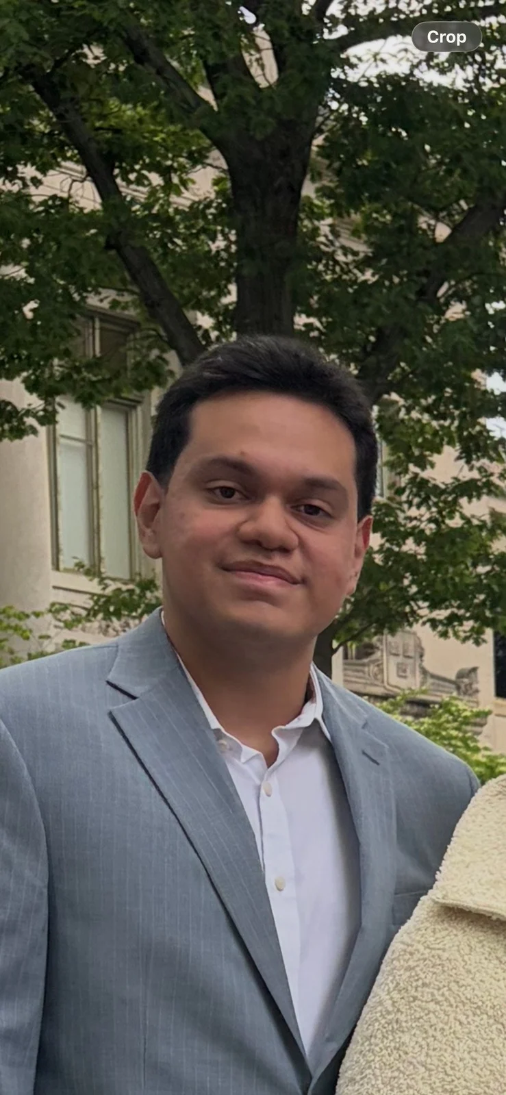 A photo of Gianni wearing formal attire in front of a tree and tan colored building