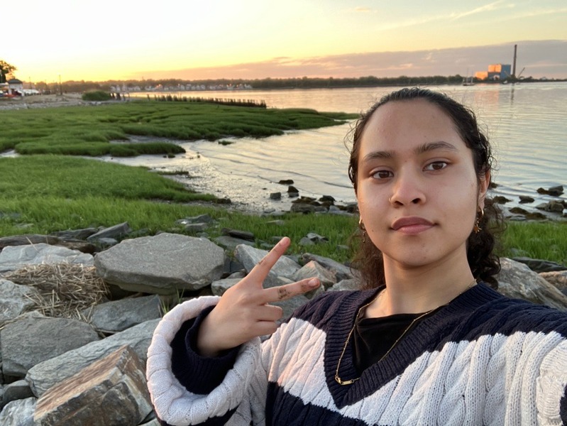 A selfie of Roshni making a "peace-sign" hand gesture from a grass and rock shore in front of a body of water.